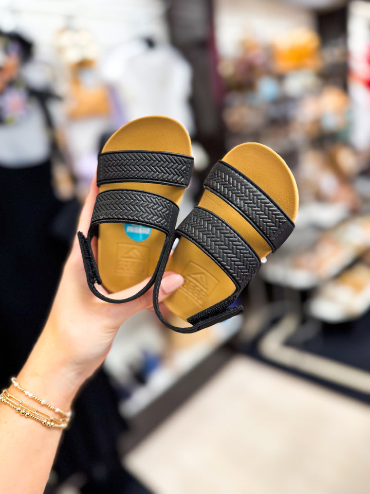 Hand holding a children's Reef Water Vista sandal with black straps and yellow insole in a store setting
