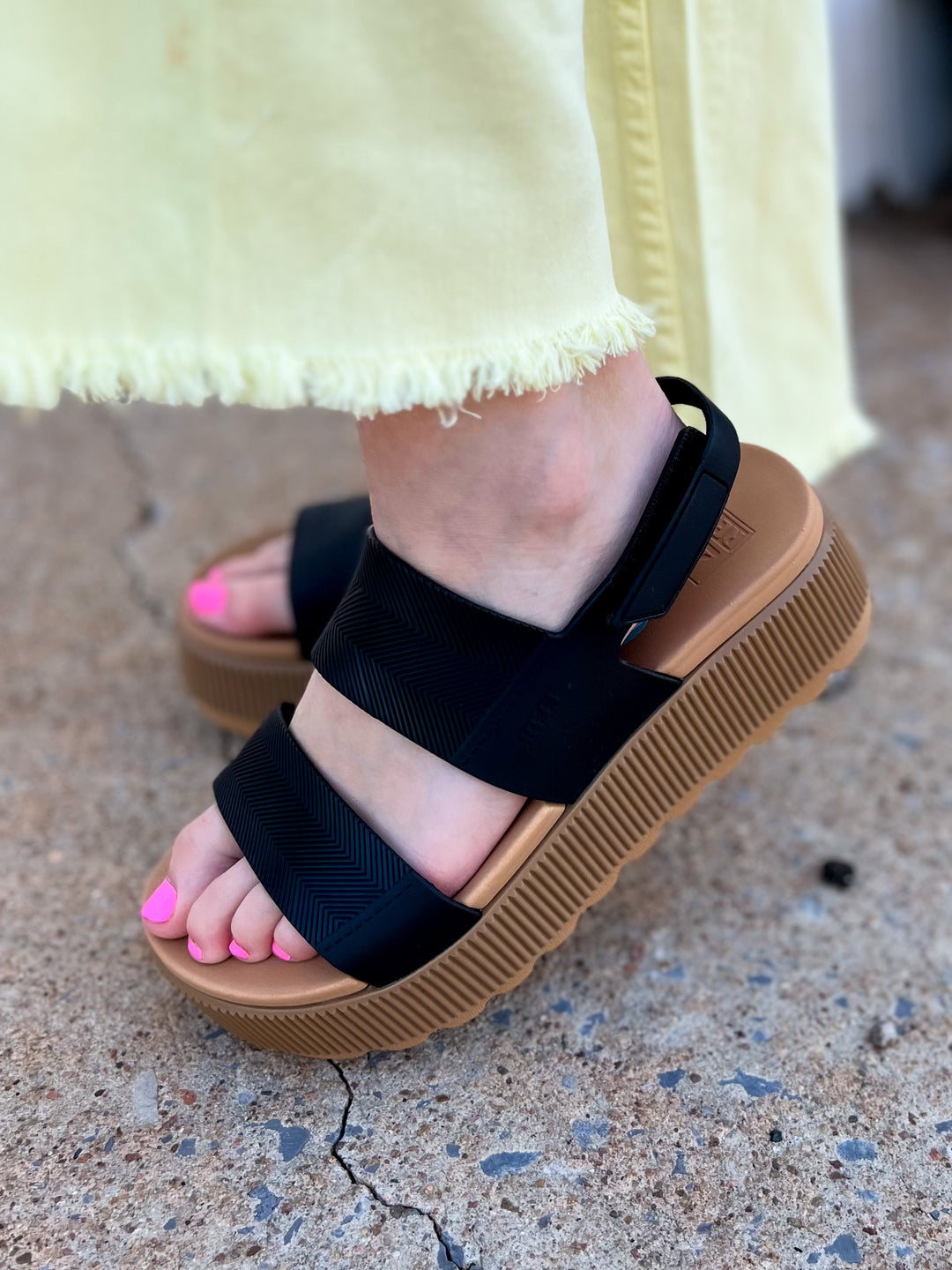 A close-up of a woman's foot wearing a Reef Higher Vista black and tan platform sandal with an adjustable velcro strap and a lightweight sole.