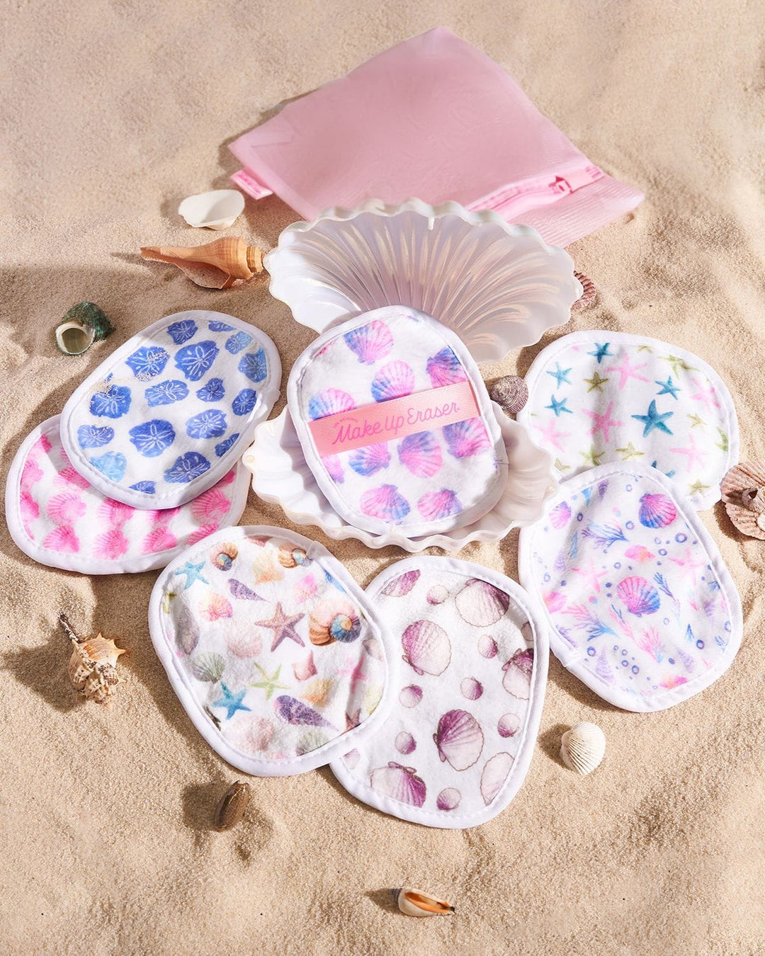 Seashell-shaped makeup erasers with ocean-themed patterns displayed on beach sand with shells