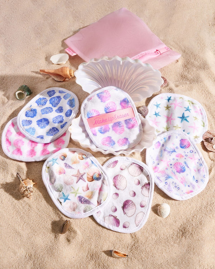 Seashell-shaped makeup erasers with ocean-themed patterns displayed on beach sand with shells