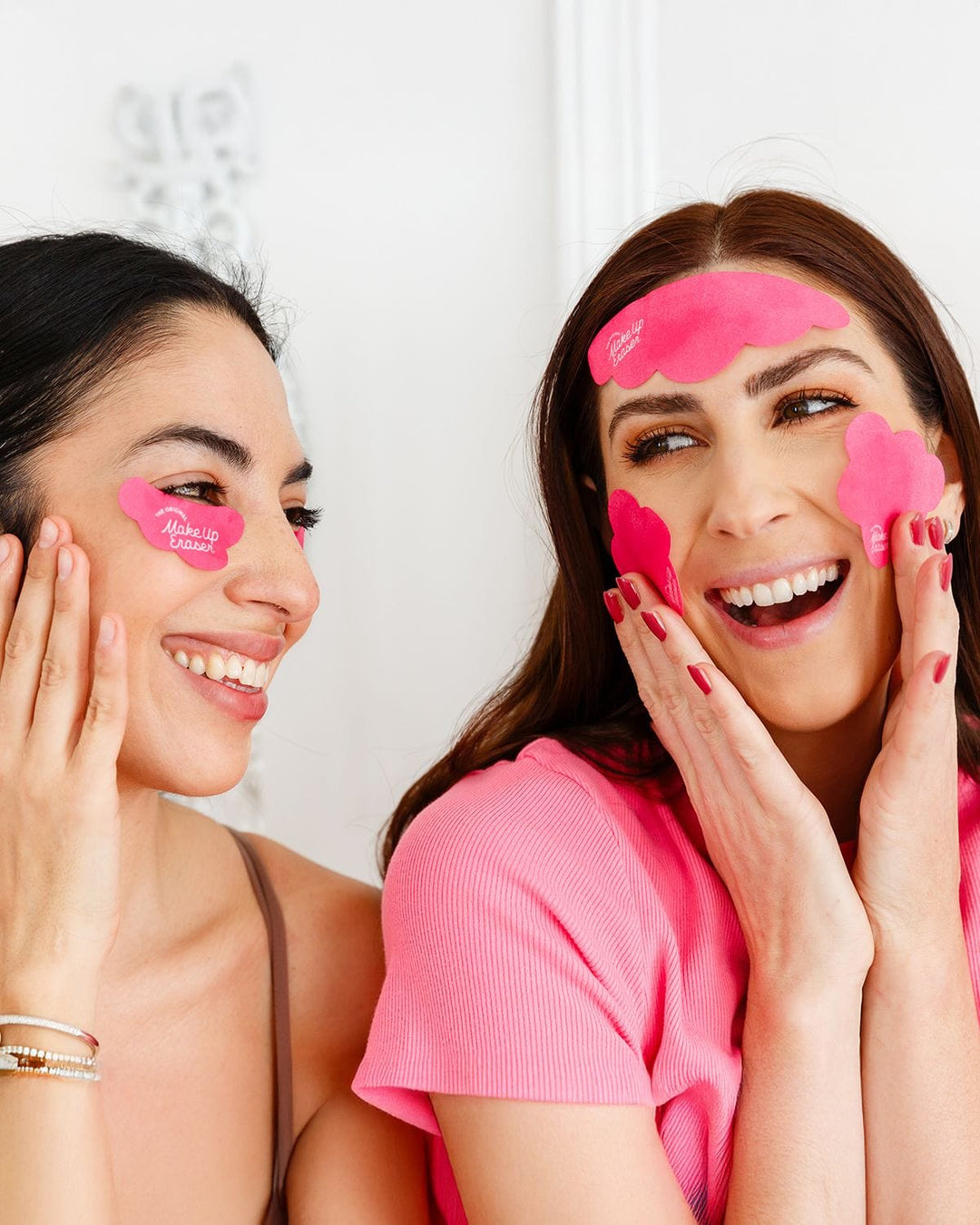 Two women wearing pink cooling under eye patches, smiling and touching their faces